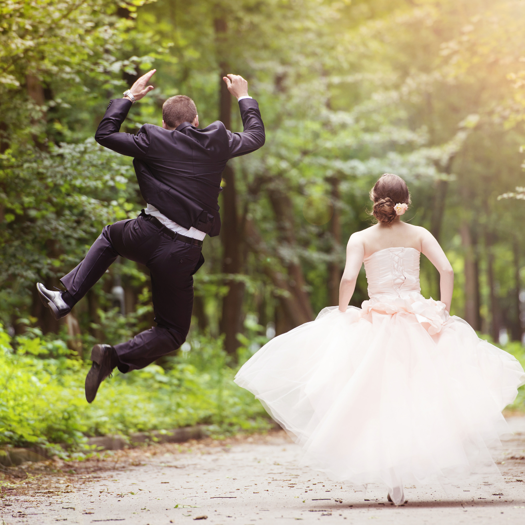 Couple côte à côte de dos marchant sur un chemin en forêt. Le marié saute. Wedding Planner Ile de France. Paris en Noces