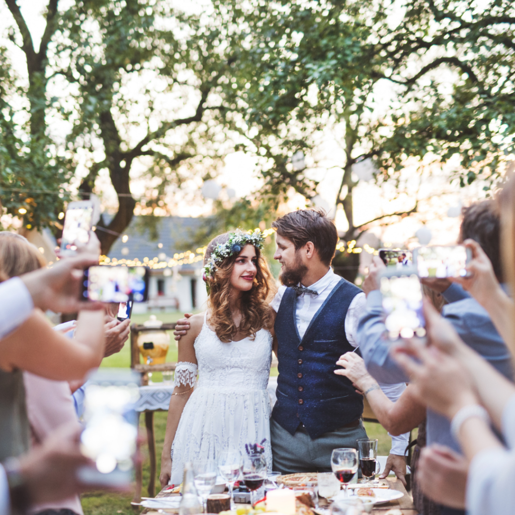 Couple de mariés dans un jardin pris en photo par leurs invités. Réussir le plan de table de son mariage. Wedding Planner Ile de France. Paris en Noces